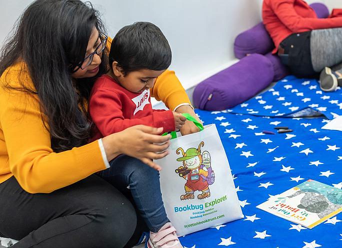 A young boy sits on a womans lap opening a Bookbug Explorer bag